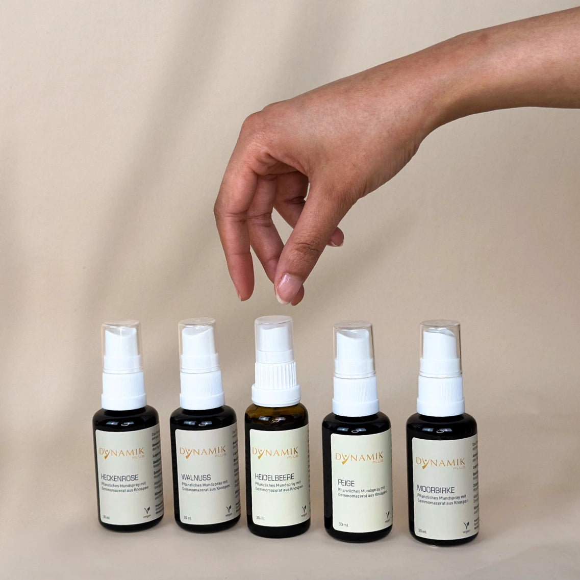 5 braune Sprühfläschchen mit weißem Sprühkopf und DynamikPlus Etikett, das mittlere wird von einer Hand gehalten